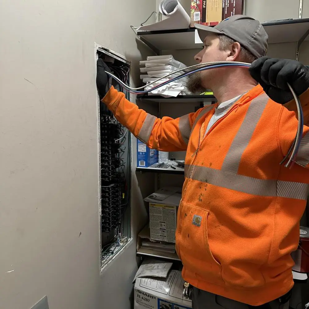 Electrician in an orange jacket working on a circuit panel, pulling wires from the wall.